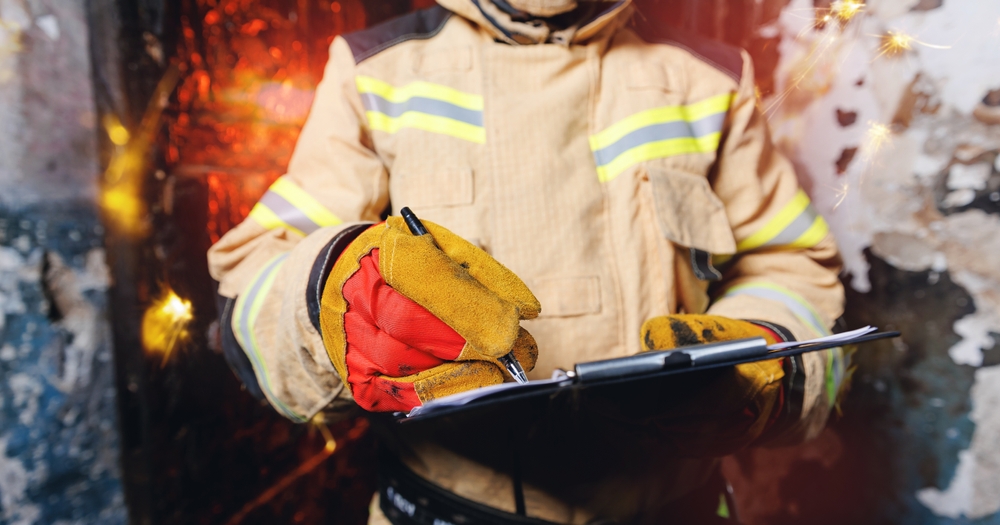 Firefighter in protective gear writing notes on a clipboard during a wildfire damage investigation